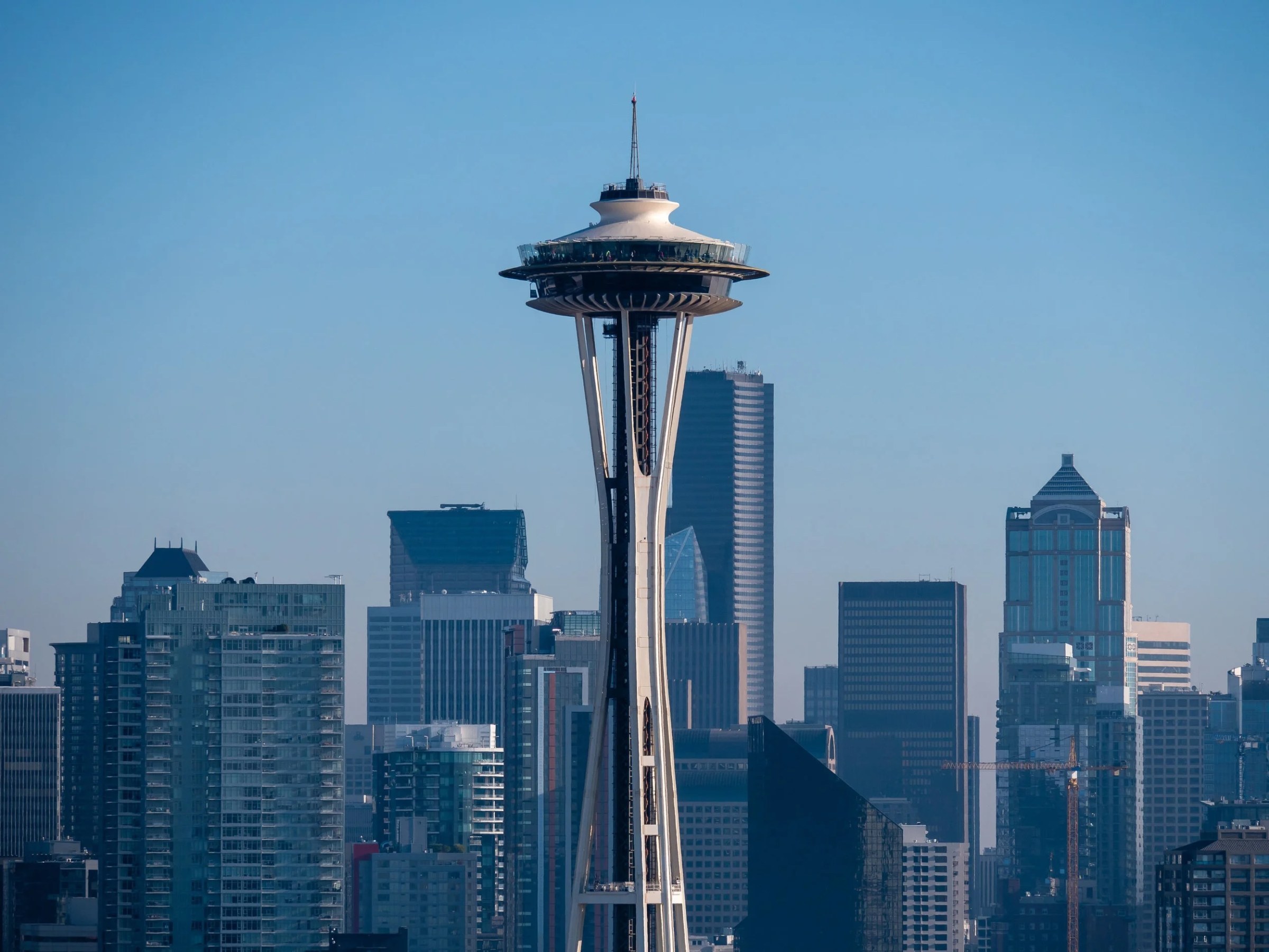 A close telephoto view of Seattle’s Space Needle, centered against a cloudless blue sky. Modern high-rises, apartment towers, and office buildings fill the foreground and background, emphasizing the landmark’s distinctive saucer-shaped observation deck and slender white supports.