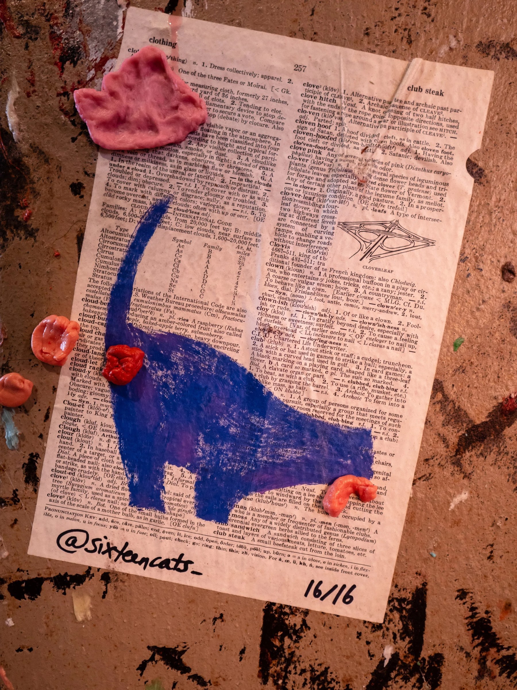 An open dictionary page, visibly worn and paint-splattered, becomes the ground for an expressive mixed-media composition. Thick swaths of deep blue and pink paint, along with several small sculpted pink and red forms, cover and interrupt the printed definitions; handwritten text at the bottom identifies the piece as “@sixteencats— 16/16.”