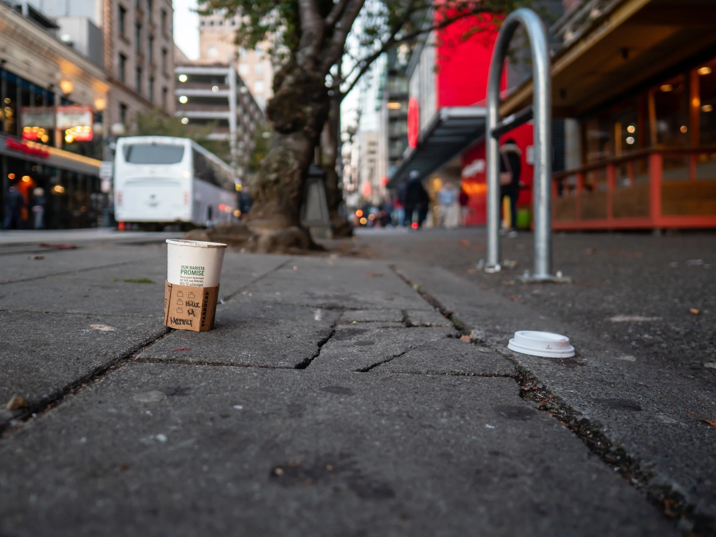 A low-angle street photograph focused on a used Starbucks paper cup standing on cracked pavement, with its plastic lid lying several feet away. The surrounding downtown scene falls softly out of focus: a white coach bus, storefronts, pedestrians, mature street trees, and a bright red building façade suggest a busy urban corridor.