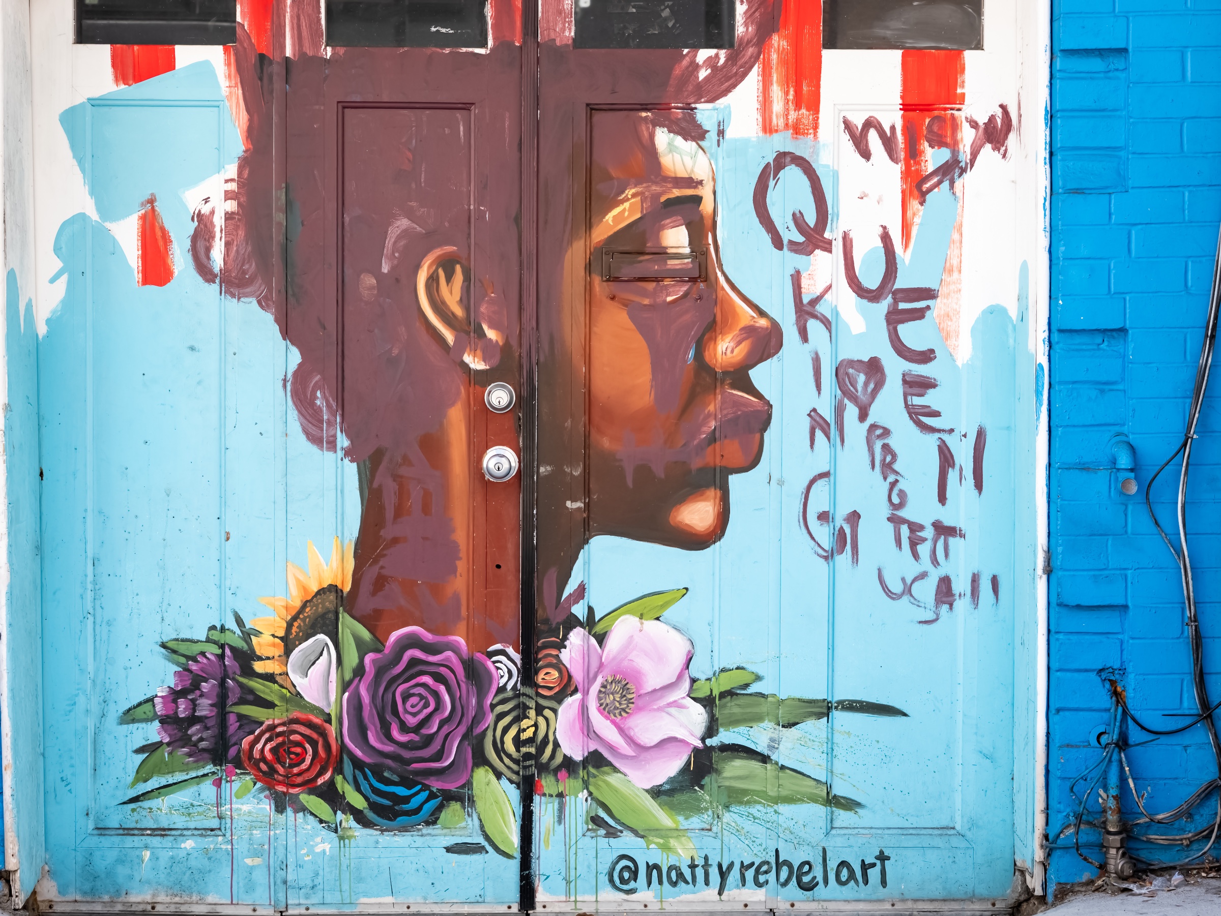 A painted mural transforms a pair of turquoise double doors into a large, contemplative portrait. The face spans both panels in warm shades of brown and copper, framed below by a vivid bouquet of roses, poppies, and sunflowers; loose handwritten marks and the artist’s handle, “@nattyrebelart,” remain visible along the right side and lower edge.