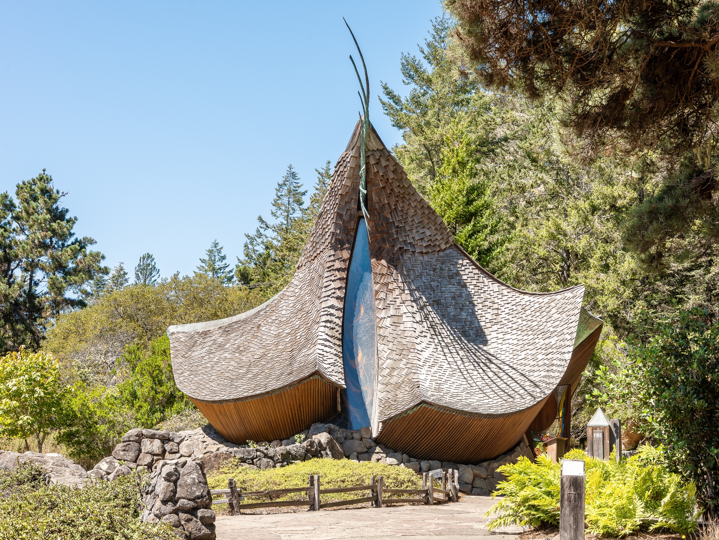 A sculptural wooden sanctuary in Sea Ranch, California, is framed by evergreens, ferns, and stone landscaping. Its sweeping cedar-shingle roof climbs to a slender central peak, while curving walls and a narrow blue-glass entrance panel give the chapel an organic, windswept presence.