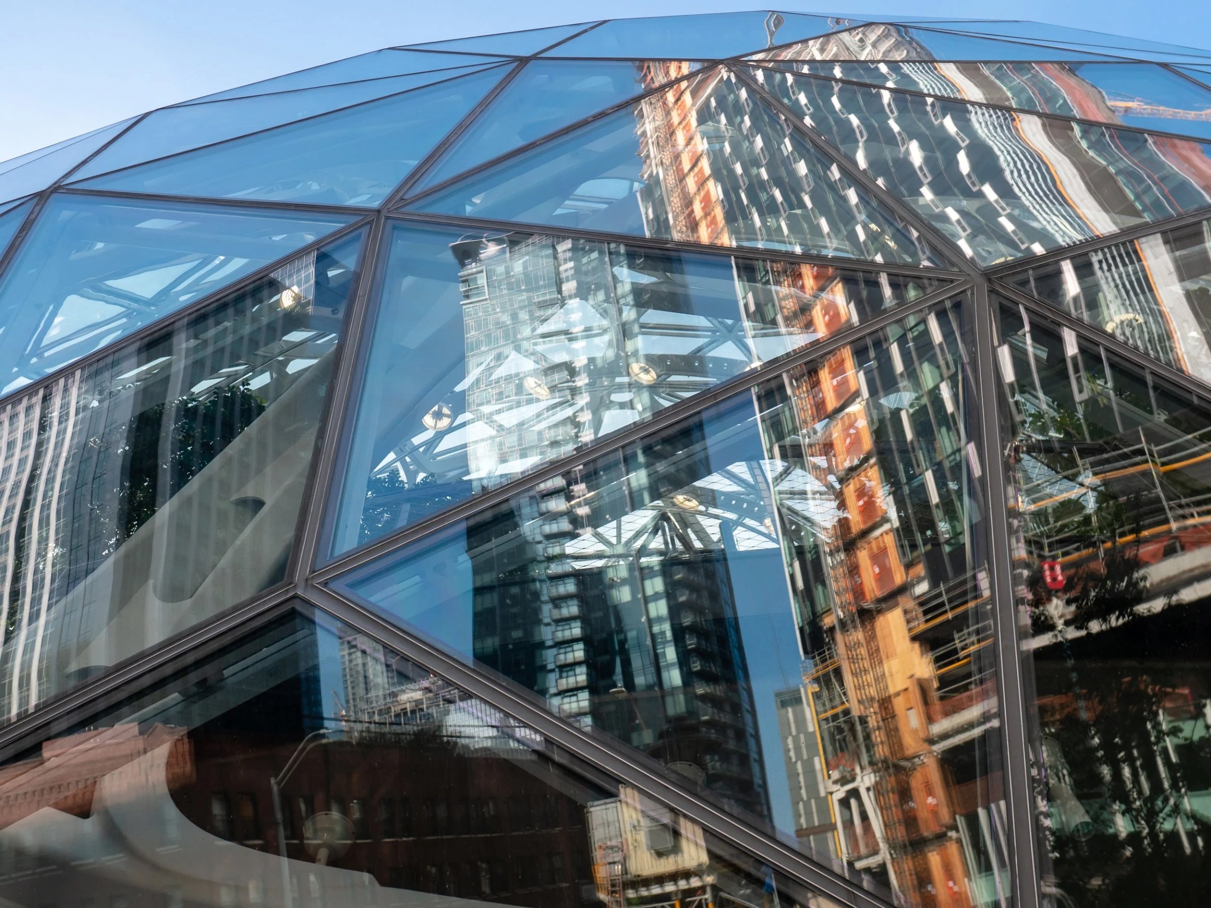 An upward-looking architectural photograph captures a modern glass-and-steel structure made of angled, triangular panes. The reflective surface layers blue sky, neighboring high-rises, construction elements, trees, and interior framework into an abstract collage, while a curving white staircase is visible behind the glass at lower left.