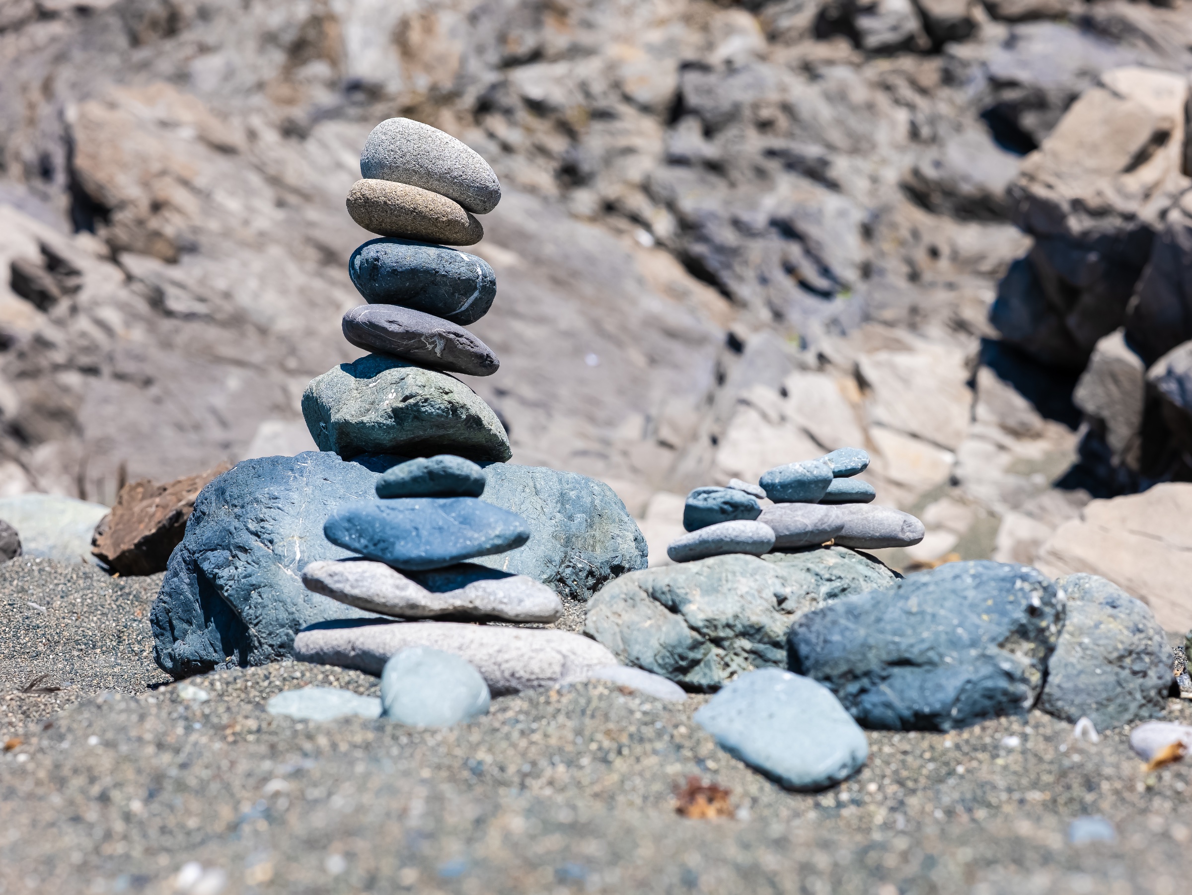 Smooth stones are carefully stacked into a delicate cairn on a rocky beach, with several smaller arrangements nearby. The close focus highlights the precarious balance and varied textures of the stones, while the weathered coastal rocks behind dissolve into a soft blur.