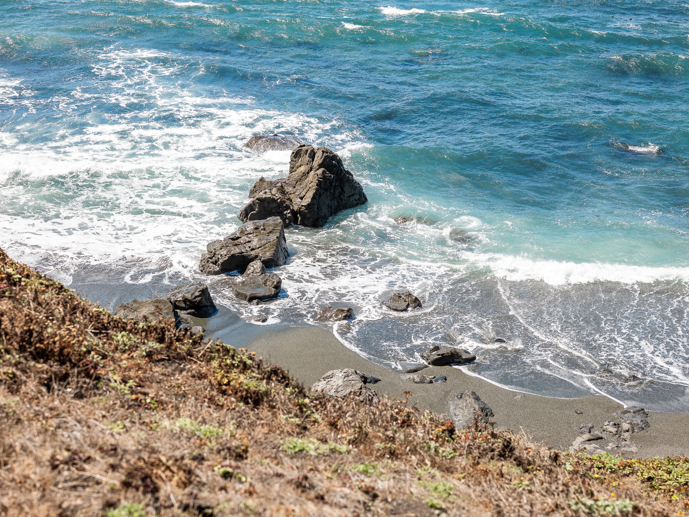 Seen from above, turquoise water curls into white foam around a cluster of dark, jagged rocks at the edge of a narrow gray-sand beach. Dry coastal grasses and low groundcover frame the foreground, while the surf traces shifting patterns across the shore below.