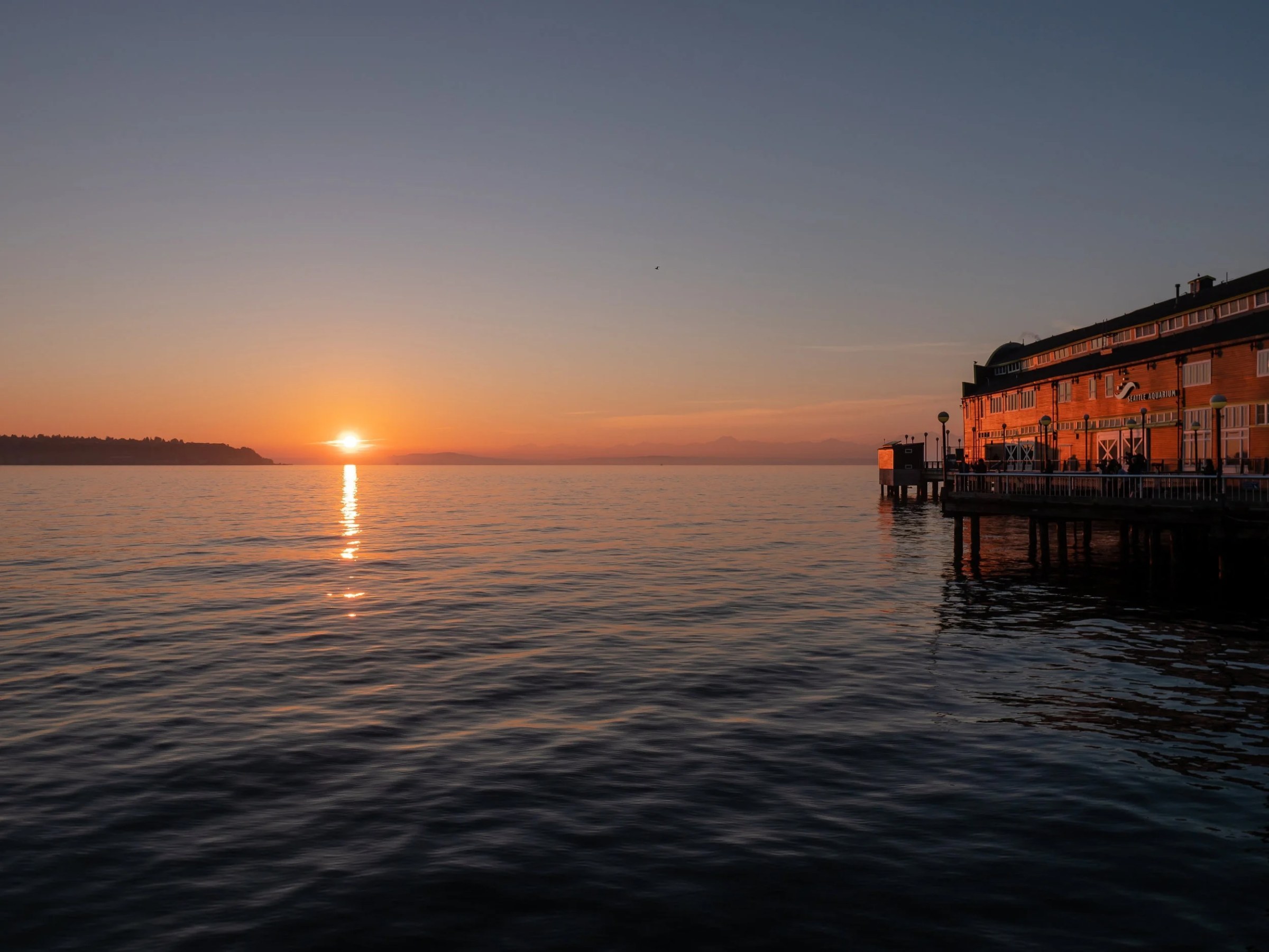 The sun sits low on the horizon beyond calm water, casting a bright golden path across Elliott Bay. At right, the Seattle Aquarium’s waterfront building and pier are warmed by the fading light, while dark, gently rippling water fills the foreground and distant shoreline silhouettes frame the sunset.