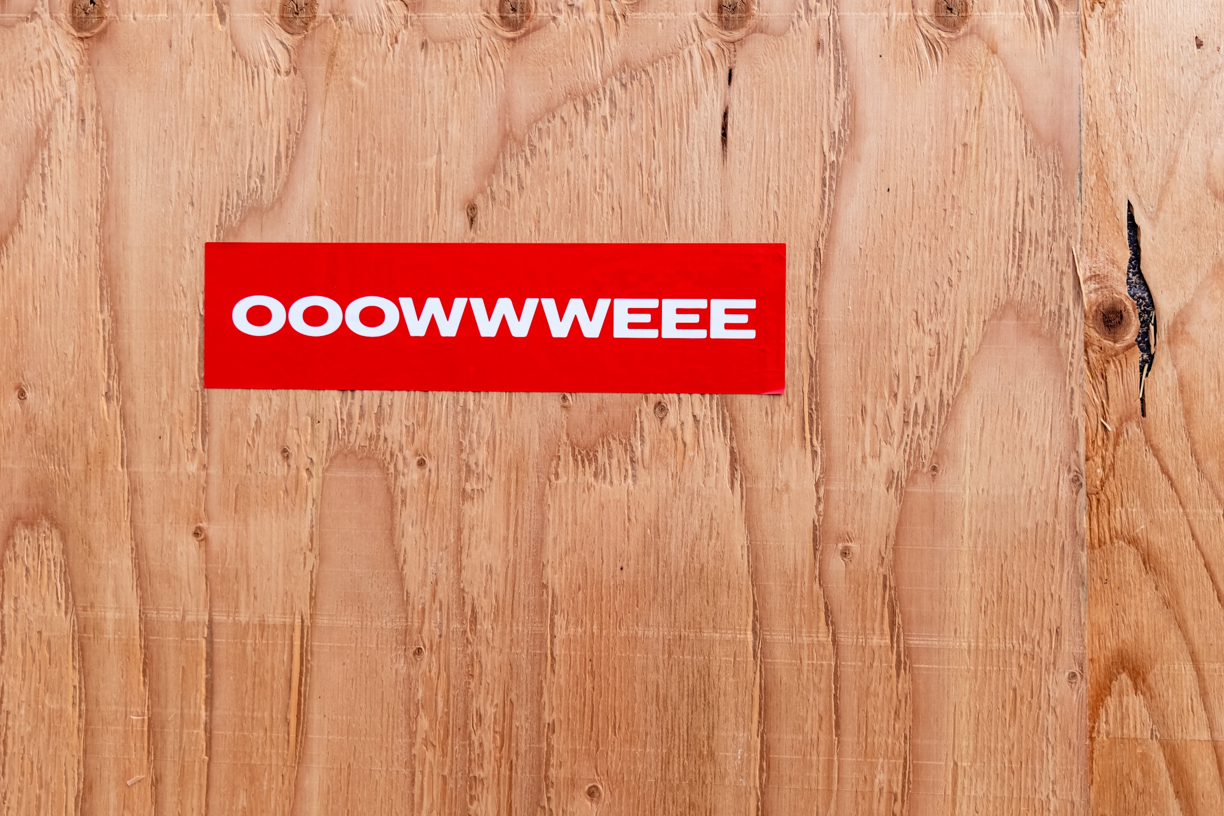 A bright red rectangular sign with the white text “OOOWWWEEE” is mounted against a wall of unfinished plywood. The raw wood grain, knots, seams, and rough opening at right create a stark, tactile contrast with the sign’s crisp color and playful typography.