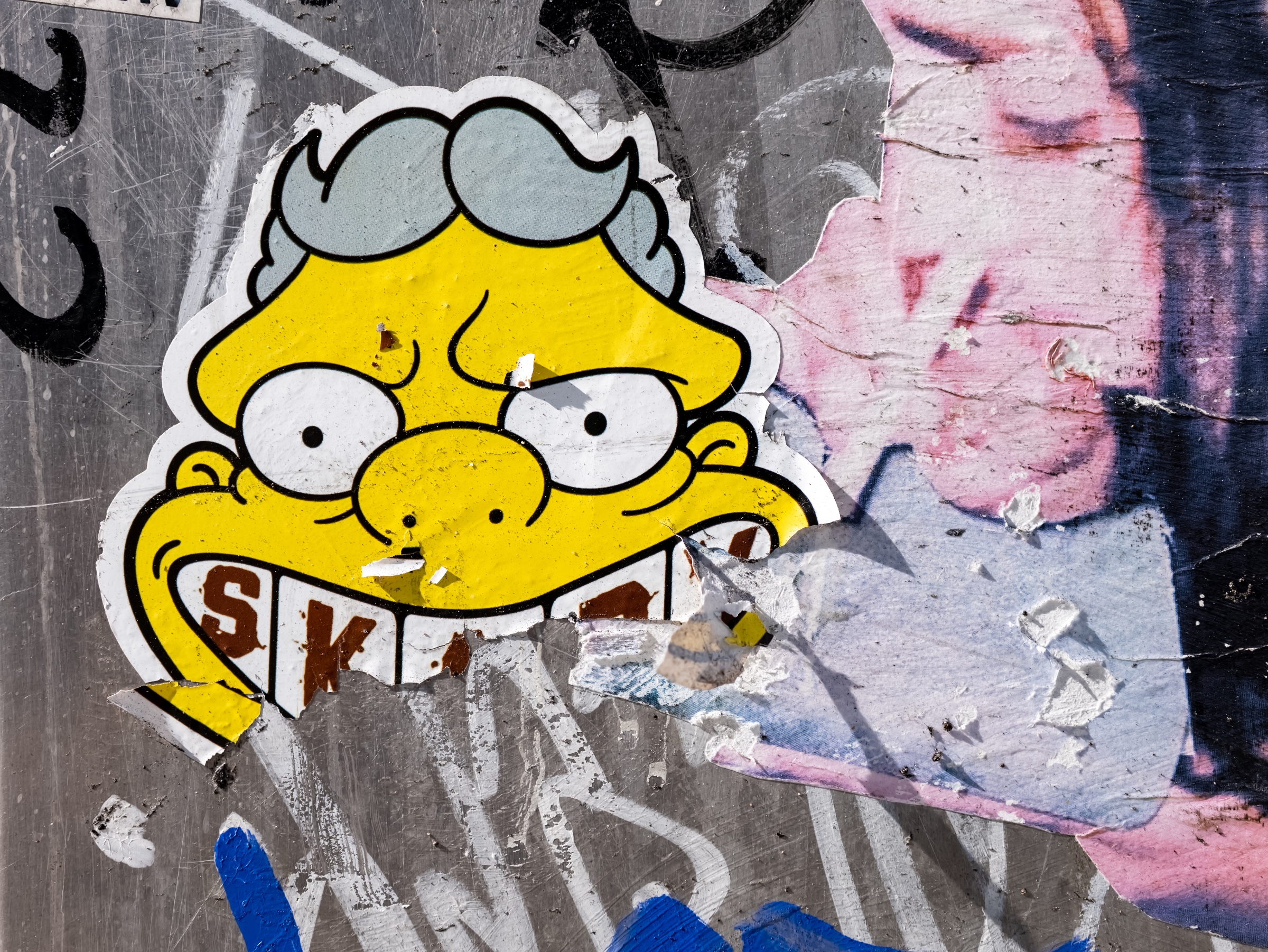 A bright yellow sticker depicting Moe Szyslak from The Simpsons—wearing gray sunglasses and a broad, toothy grin—has been pasted onto a battered wall of overlapping posters, paint, and graffiti. Torn paper, scratches, adhesive remnants, and fragments of pink and black imagery turn the surface into a dense, weathered urban collage.
