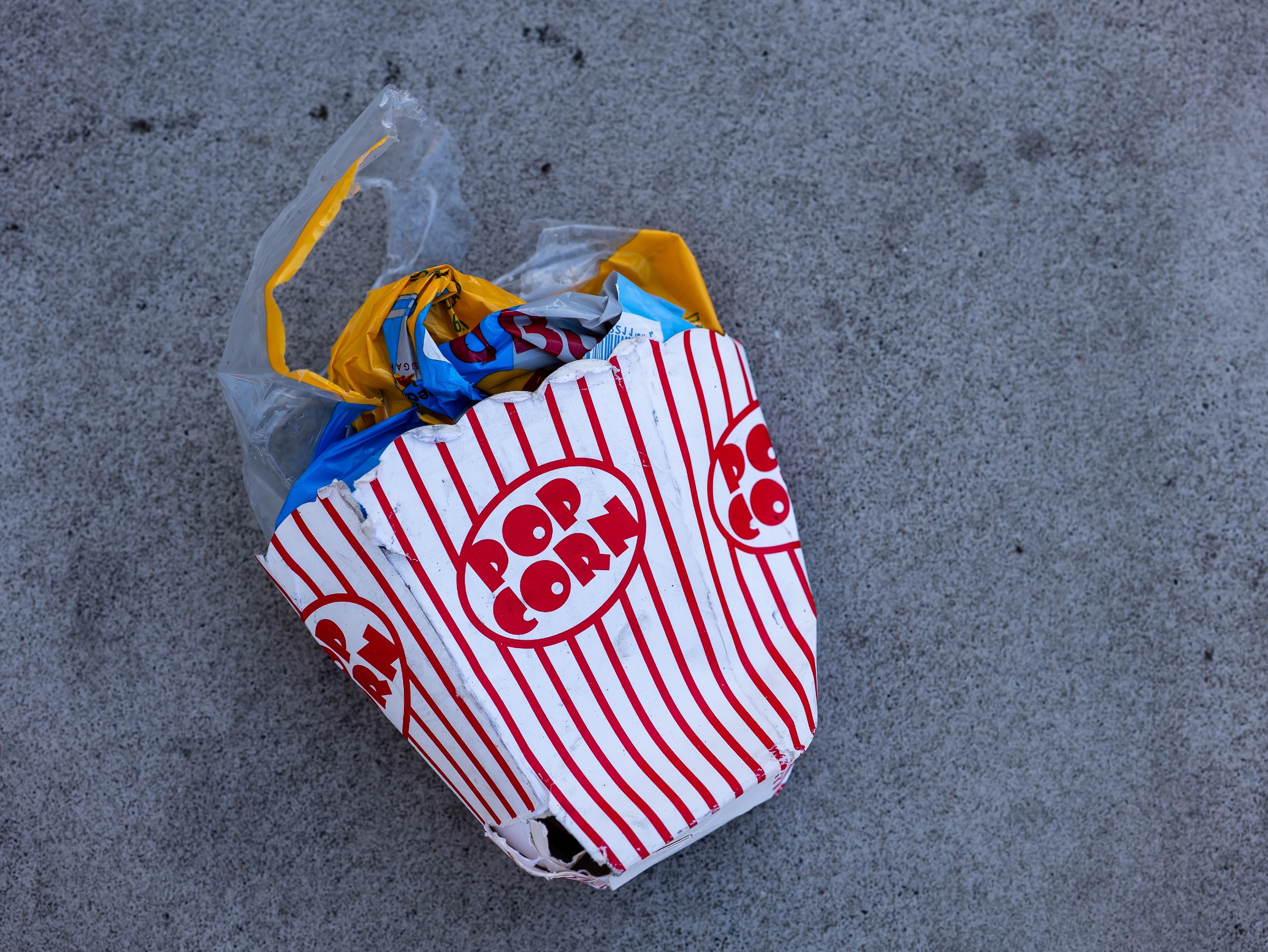 A crushed striped popcorn box and torn plastic snack wrappers lie on a gray concrete surface. The bright red, yellow, and blue packaging contrasts sharply with the muted pavement, turning a small piece of street litter into a study of color, consumption, and aftermath.